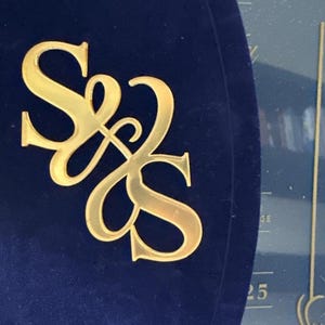 May include: A navy blue, velvet-like surface with a gold-colored monogram of the letters "S" and "Z" intertwined. The monogram is shiny and reflective. The background includes a portion of a calendar with the number "25" visible.