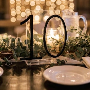 May include: A table setting with a clear acrylic table number 10. The number is black and has a modern font. The table is decorated with greenery, white plates, and a water pitcher. Soft lights are in the background.