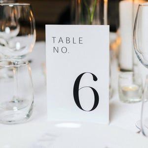 May include: White card with black text that reads "TABLE NO. 6". The card is sitting on a white tablecloth with two clear glass goblets and a small candle holder.