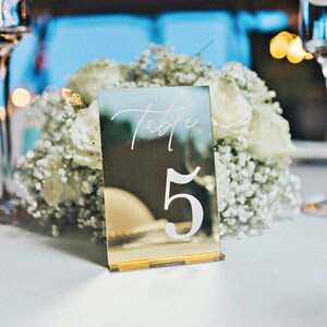 Puede incluir: Una tarjeta de número de mesa de espejo dorado con la palabra "Table" y el número "5" escritos en blanco. La tarjeta está sobre un mantel blanco con un ramo de flores blancas en el fondo.