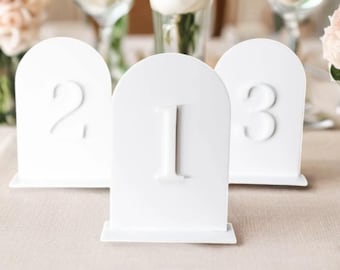 May include: Three white table number signs with rounded tops and a base. The numbers 1, 2, and 3 are embossed on the signs.