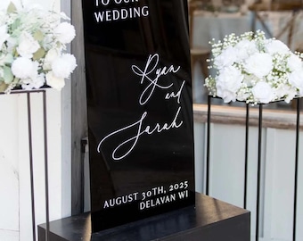Puede incluir: Un letrero de bienvenida de boda de acrílico negro con letras blancas. El letrero dice "Bienvenida a nuestra boda" y presenta los nombres "Ryan y Sarah" junto con la fecha y el lugar. El letrero se muestra en un pedestal negro.