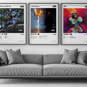 May include: Three framed album art prints hang above a gray sofa. The prints feature colorful artwork and text, including the artist's name and album titles. A potted plant and a small side table with decor complete the room.