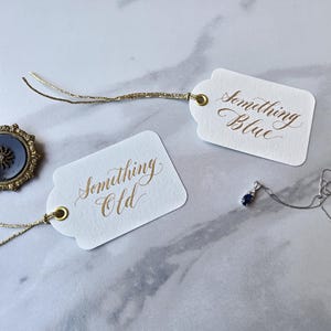May include: Two white rectangular paper tags with gold metallic strings and brass eyelets. One tag features the gold calligraphy text Something Old, and the other features Something Blue. A dark circular brooch and a silver necklace are included.