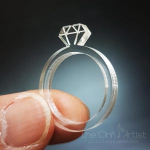 May include: A transparent ring with a diamond-shaped top, held against a dark background. The ring has a double band design and a geometric diamond shape. The ring is made of a clear material, giving it a delicate appearance.