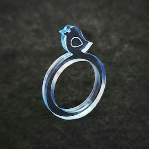 May include: A clear acrylic ring featuring a bird design. The bird is dark blue with a white outline and a small white circle. The ring is light blue and has a layered design.