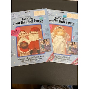 Puede incluir: Dos paquetes de caras de muñecas termoadhesivas "Dolly Dear". Los paquetes presentan imágenes de muñecas, incluyendo un Papá Noel y un ángel. El texto en los paquetes incluye "Full Color Iron-On Doll Faces" e instrucciones para hacer muñecas trapeadoras.