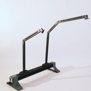 May include: A dark grey, adjustable desk lamp with two articulated arms. The lamp has a rectangular base and two upright supports. The arms extend from the supports and have small, adjustable heads. The lamp is designed for task lighting.