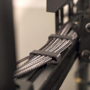 May include: A black cable management clip securing a bundle of braided black and grey cables along a black metal frame. The clip is rectangular with rounded edges, holding the textured cables in place against the structural support beam.