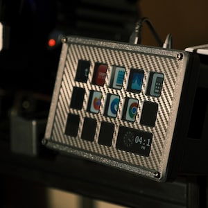 May include: A carbon fiber-textured control panel with multiple illuminated buttons and a digital clock displaying 04:11 PM. The panel is mounted on a black surface, likely part of a simulator or gaming setup.