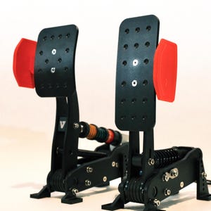 May include: A pair of black racing pedals with red accents. The pedals have a perforated foot plate and are mounted on a black frame with springs and various coloured components. The background is plain white.