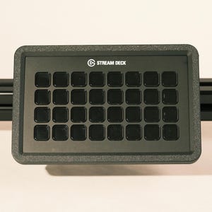 May include: A black Stream Deck with 32 square buttons. The device has a textured frame and the words "STREAM DECK" are printed in white. The device is mounted on a black rail.
