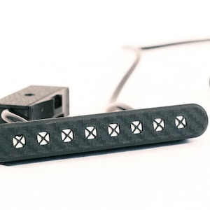 May include: A black carbon fiber guitar pickup featuring a long, rectangular housing with eight individual pole pieces arranged in a single row. The component includes a small black mounting block and thin gray wiring extending from the side.