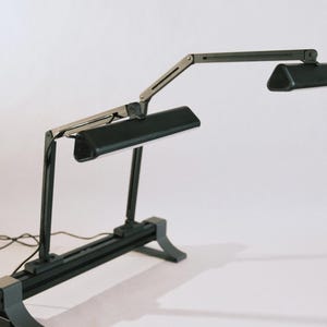 Puede incluir: Una lámpara de escritorio negra con dos cabezales de luz ajustables. La lámpara tiene una base rectangular y brazos articulados para un posicionamiento flexible. Los cabezales de luz son rectangulares e inclinados. La lámpara está diseñada para iluminación de tareas.
