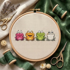 Puede incluir: Un bastidor de bordado de punto de cruz con cuatro ranas de dibujos animados en rosa, naranja, verde y blanco. Las ranas están cosidas en una tela beige, colocadas en un bastidor de madera. La imagen incluye tijeras doradas, carretes de hilo verde y flores secas.