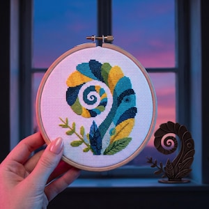 May include: A circular wooden embroidery hoop containing a cross-stitch pattern of a stylized chameleon. The design features a curled tail and body in shades of yellow, teal, dark blue, and green, set against a white fabric background.