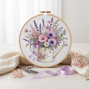 Peut inclure: Un tambour à broder circulaire en bois contenant un motif au point de croix représentant un arrosoir blanc rempli d'un bouquet de fleurs violettes, roses et blanches, incluant de la lavande et des cosmos, orné d'un ruban violet clair.
