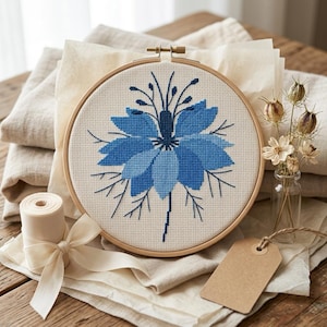 May include: A completed cross-stitch embroidery of a blue flower in a wooden hoop. The flower has layered petals in varying shades of blue. The embroidery is on a cream-colored fabric, with a wooden hoop. The background includes folded fabric, a ribbon, and a small glass vase.