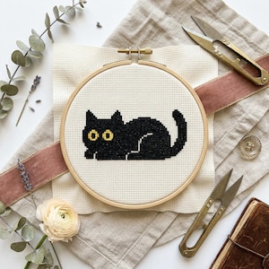 Peut inclure: Broderie au point de croix d'un chat noir aux yeux jaunes dans un cercle en bois. L'image comprend des ciseaux, un ruban rose et des éléments floraux. Le fond est un tissu crème.