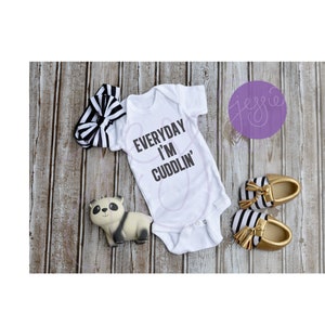May include: White baby onesie with black text that reads "EVERYDAY I'M CUDDLIN'". The onesie is laying on a wooden surface with a black and white headband, a panda toy, and black and white striped baby shoes with gold accents.