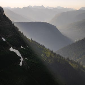 May include: A scenic landscape photograph of a mountain range. The image features layers of mountains fading into the distance, with a foreground of a steep, green hillside dotted with trees and patches of snow. Sunlight streams through the valley.