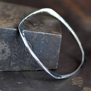 May include: A silver hammered bangle bracelet with a square shape. The bracelet is sitting on a textured metal surface.