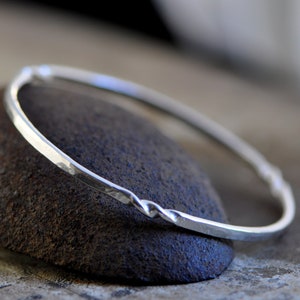 May include: A silver bangle bracelet with a hammered finish and a twisted design. The bracelet is resting on a dark grey rock.