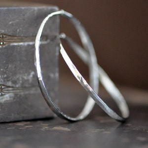 May include: A pair of silver hoop earrings with a hammered texture. The hoops are large and have a slightly oval shape.