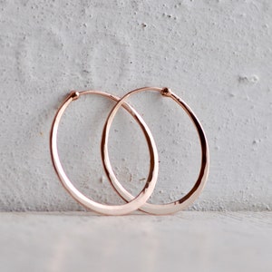 May include: A pair of rose gold hoop earrings with a subtle hammered texture. The circular earrings feature a thin, polished metal band that catches the light, creating a reflective surface. The design is simple, consisting of a continuous wire loop.