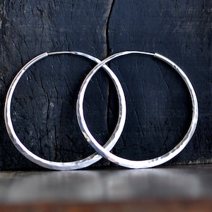 May include: A pair of large, silver hoop earrings with a hammered texture. The earrings are thick and have a slightly oval shape.