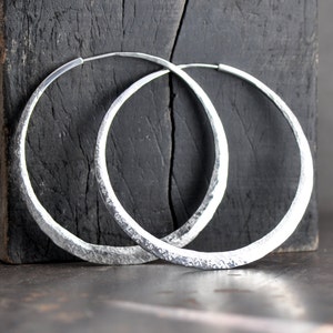 May include: A pair of large, silver hoop earrings with a hammered texture. The earrings are made of metal and have a smooth, polished finish.