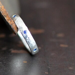 May include: A hammered silver ring with three small, round, blue gemstones. The ring is set against a dark brown background, with a piece of metal in the upper left corner.