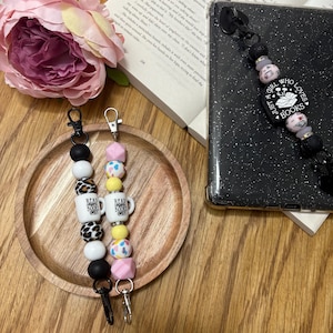 May include: Two beaded keychains with black, white, and patterned beads. One keychain has a small mug charm that says "Read Books". A phone strap with black beads and a charm that says "A Girl Who Loves Books" is also visible.