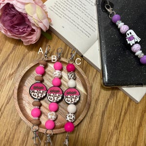 May include: Three pink, brown, and white beaded phone charms with a leopard print design and the word "mama" on a round charm. The charms are hanging from silver key rings.