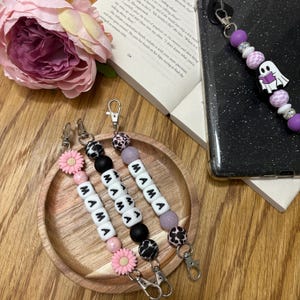 May include: Three beaded keychains with the word "MAMA" spelled out in white letter beads. One keychain has pink daisy and pink beads, another has black beads, and the third has lavender beads. A phone with a purple beaded strap is also visible.