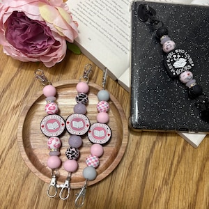 May include: Three beaded keychains with lobster claw clasps. The keychains feature pink, grey, and black beads, with a circular charm that reads "BAD BITCH BOOK CLUB." A phone with a black beaded strap that says "GIRL WHO LOVES BOOKS" is also visible.