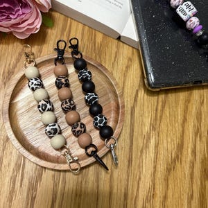 May include: Three phone charms with black, brown, and beige beads with a leopard print pattern. Each charm has a gold or black metal clasp.