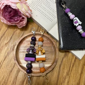 May include: Two purple and orange Halloween phone charms with dragon and ghost figures. The charms are made of beads and have a key ring attached.