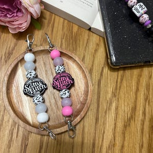 May include: Two beaded keychains with the text "Salty Bitch Club" on black and pink circular charms. The keychains feature white, gray, pink, and patterned beads. One keychain is on a wooden tray.
