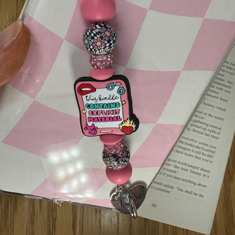Op de afbeelding: Een roze kralen sleutelhanger met een bedel met de tekst "This Kindle Contains Explicit Material". De sleutelhanger heeft decoratieve kralen in roze, zwart en wit, en een hartvormige bedel.