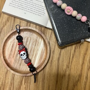May include: A phone charm with a skull design, red bow, and black beads. The charm is attached to a black clasp. The charm is on a wooden plate. The beads are in various sizes.