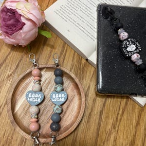 May include: Two gray and pink beaded phone charms with a heart-shaped charm that says "Book Lover" on each. The charms are hanging from silver key rings. The charms are on a wooden surface.