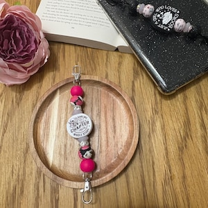 May include: Pink and white beaded phone charm with a round charm that says "You're a darling, just a chapter in the whole story".