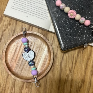 May include: A beaded keychain with a heart-shaped charm featuring a skeletal design. The keychain includes purple, teal, and patterned beads, and a silver clasp. The background includes a wooden tray, a book, and a phone case.