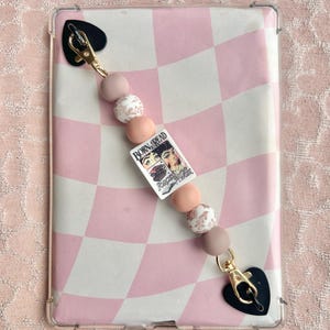 May include: A clear phone case featuring a pink and white chequered pattern. A beaded strap with pink, white, and mauve beads is attached. The strap includes a rectangular charm with the text "BORN BREAD FORGET ME NOT". Black heart-shaped clasps complete the design.