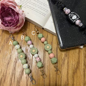 May include: Three green and pink beaded keychains with floral accents and gold clasps. The keychains are labelled A, B, and C. The text "Girl Who Loves Just A Book" is visible on a black phone case.