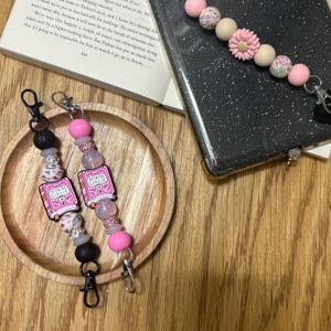 May include: Two beaded lanyards with black, pink, and leopard print beads. Each lanyard has a charm that says "Bookish Girlie" and a lobster clasp. A phone case with a beaded strap is also visible.