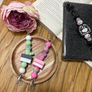 Könnte beinhalten: Zwei Handy-Anhänger im Buchwurm-Design mit grünen und rosa Perlen, einem Buch-Anhänger und einem silbernen Schlüsselring. Die Anhänger befinden sich auf einer Holzoberfläche. Der Text auf dem Anhänger lautet "Just a girl who loves books".