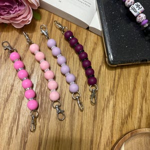 May include: Four beaded phone charms in shades of pink, purple, and black. Each charm features a series of round beads and a silver-colored clasp. One charm has white beads with black text. The charms are displayed on a wooden surface.