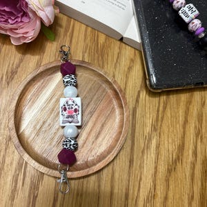 May include: A white and purple phone charm with a white bead, a white and black leopard print bead, a burgundy bead, a white bead with a book charm that says "Give us a break" and a white and black leopard print bead. The charm is attached to a silver key ring.